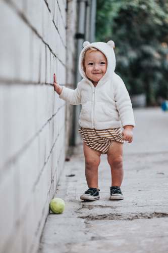 smiling baby girl in white hoodie and brown and white polka dots skirt holding green tennis ball grey